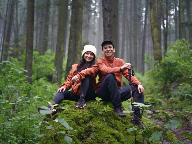 a couple both wearing red tops and blue trousers sit on a mossy boulder in a dense green forest during a walk the man holds a walking stick
