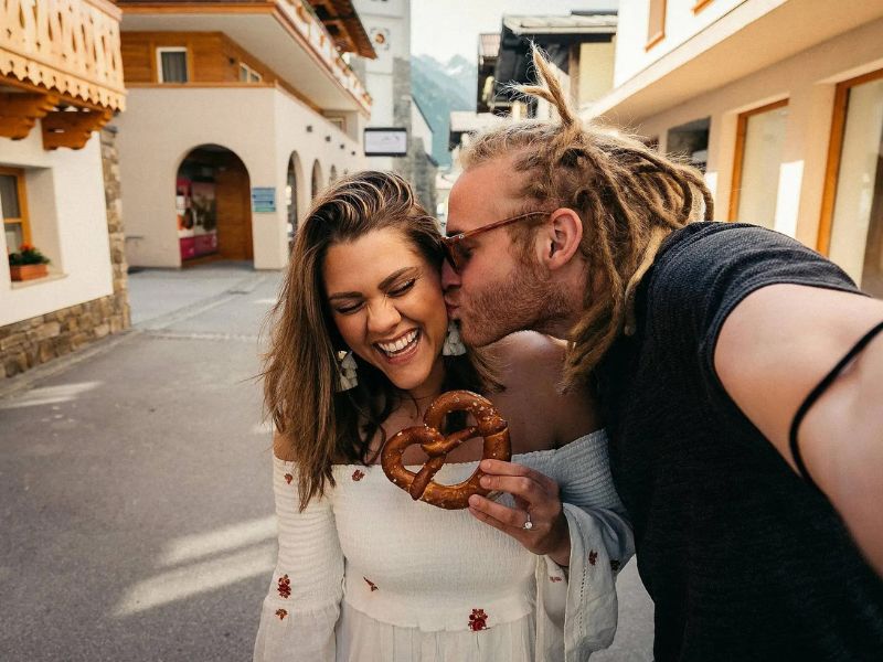 alternative travel companions embrace for a selfie on a city break; woman holds a bagel while the man takes the click